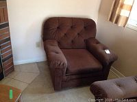 Front view of the brown overstuffed armchair positioned near a wall and tiled floor, showing tufted backrest and cushioned armrests.
