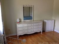 Wide angle photo of entire white dresser with nine drawers and mirror on top, showing its size and shape in a room setting.