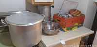 Overview of large aluminum stockpot with lid, metal pail, stainless steel colander, and cardboard box with assorted kitchen items on kitchen counter.