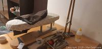 Photo showing a compact ironing board with padding and cover, metal legs, measuring sticks leaning against the wall, bottle of pressing aid, metal tray with various tailoring tools and spools of thread.