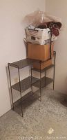 Metal shelving unit with three mesh wire shelves, placed against a white wall on a concrete floor, with two boxes on top containing fabric remnants.