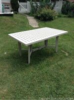 Full side angle of the white plastic patio table on grass outdoors showing slatted top and legs.
