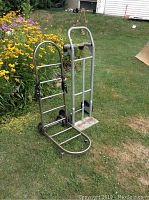 Two adjustable metal dollies side by side on grass near flowers, showing condition and structure.