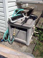 Plastic hose storage box with garden hose and Yardworks oscillating sprinkler sitting on top, outdoor setting beside wall