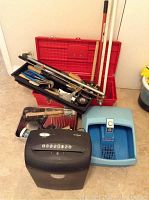 Photo of red plastic toolbox open showing various hand tools in black removable tray inside. Two long handled tools leaning against the toolbox. Blue foot massager and black Omnitech paper shredder in front on floor.