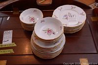 Photo showing stacks of floral porcelain plates and bowls with gold trim on a wooden surface.