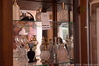 Two glass shelves in a wooden cabinet holding 11 vintage glass perfume bottles of various designs and shapes made from clear and frosted glass with gold-tone spray pumps and decorative stoppers.