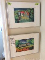 Two framed watercolour paintings hanging; one depicting houses with red roofs, the other houses with dark roofs and greenery. Both have white frames and mats.
