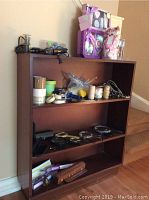 Full view of brown wooden bookcase with multiple shelves holding magnifying glasses, rolls of tape and twine, some other craft items and personal care gift sets on top.