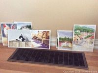 Seven watercolor paintings displayed on the floor showing a variety of subjects including animals, landscapes, and village scenes.
