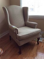 Side angle view of Queen Anne wingback chair upholstered in beige patterned fabric, showing wooden cabriole front leg and overall condition.