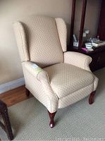 Frontal view of the beige wingback recliner chair with patterned fabric upholstery and rolled arms, showing wooden legs.