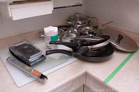 Overview of several Palermo and Lagostina pots and pans stacked together with some glass lids, plus a black-handled frying pan, and a metal strainer on the side on kitchen counter.