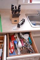 Knife block with 12 Cuisinart steak knives and manual knife sharpener next to kitchen sink, with drawer of mixed cooking utensils below.