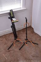 Two black metal guitar stands with orange rubber protective padding on the arms, one folded adjustable Apex sheet music stand beside them on brown carpet near white wall and window.