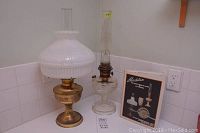 Photo of two vintage Aladdin oil lamps and hardcover book on a white tiled surface showing lamp condition and book cover.