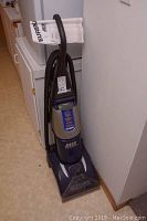 Upright blue Eureka carpet cleaner with hose and handle leaning against a wall, showing overall form and color.