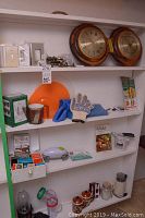 Photo of 4 white shelves filled with various household items including a Magic Bullet blender base with two cups, wood wall clock/barometer, gloves, decks of cards, books, plates and light switch faceplates.