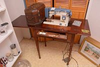 Full view of the sewing machine mounted on the wood table with the brown studded box to the left side. The drawer of the table is partially visible with sewing accessories inside.