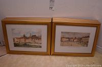 Two gold framed prints leaning against wall showing village and architectural scenes
