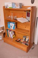 Bookshelf with three shelves containing various items including books, glassware, and boxed items, along with a bottom drawer with brass knobs. Taken to show the overall condition and details of the bookshelf.