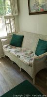 Full view of the white wicker three cushion sofa with floral seat cushions and two green throw pillows, positioned against wall with window and wood shutters.
