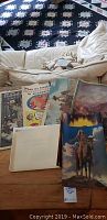 Image showing vintage western art prints, vintage newspaper ads including strawberry ad, and autograph sheet placed on wooden surface in front of beige couch with quilted pillow
