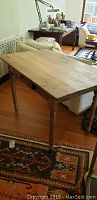 Full view of the antique pine table showing flat rectangular top with stains and four turned legs on a wood floor next to rugs and furniture.