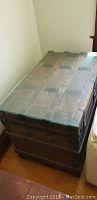 Full view of the wooden trunk showing the glass top surface, dark metal strap hinges and overall structure.