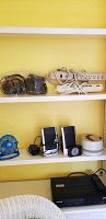 Shelf showing power strips, assorted headphones (3 pairs), blue USB fan, two black speakers, a small clock, and white cylindrical sound soother device, along with a black Memories VHS player below.