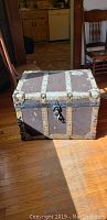 Full view of the vintage trunk placed on a wooden floor, showing its rectangular shape and metal hardware.