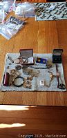 Overview of the costume jewelry lot laid out on a textured cloth showing all watches, lockets, cufflinks, earrings, and small brooch.