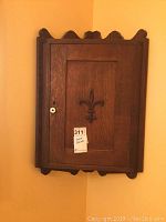 Front view of wooden hanging corner cabinet showing door with fleur de lis applique and scalloped edges.