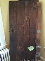 Full frontal view of antique wooden door showing surface wear, door handle, and lock hardware, leaning against interior wall near window and radiator.