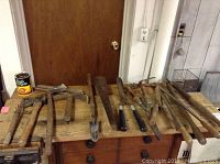 Photo showing full assortment of hand tools including axe, pick axes, shovel, cutters, shears, handles, dowels, and cage on wooden workbench surface.
