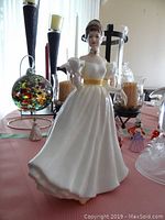 Porcelain Royal Doulton figurine of a woman in white dress with yellow sash, surrounded by glass candle holders and other glass items.