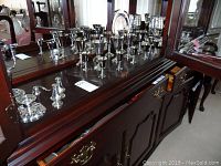 Wide view of many silver plated items including multiple tall candle holders, creamer and sugar set, glasses, tray and water jug all displayed on wooden cabinet.