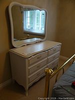 Photo of six-drawer white dresser with gold accents and curved edges, showing front and top surface, placed next to a bed in a bedroom.