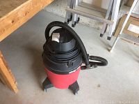 Shop Vac unit on wheels showing red body and black top part with attached hose coiled.