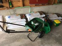 Full side view of Scotts push seed spreader with handle, wheels, and green hopper shown among bags of topsoil.