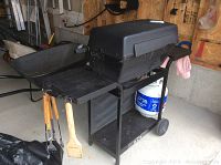 Side view of black Broil Mate propane BBQ grill with side shelves, propane tank under side shelf, and wooden handle BBQ tools hanging on hooks attached to grill.