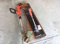 Black+Decker cordless weed eater lying on floor next to its box showing full length of tool.