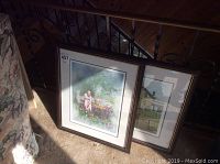 Two framed prints by Roger Witmer leaning against a wall, one showing a woman tending a garden, the other a house with a wagon in front.