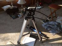 Aluminum adjustable tripod with alt-azimuth mount shown assembled and standing on carpeted floor. Various household items visible in background.