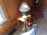 Photo of a small wooden table with a green ceramic base lamp on top and a white candle bowl, along with several picture frames and prints on the carpet.