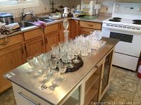 Crystal and glass tableware on metal island counter including goblets, tumblers, decanter, candy dishes on wooden-serving tray.
