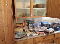 Kitchen countertop and cabinet with silverware tray, serving dishes, crock pot, food processor, cookbooks and aprons all visible