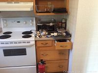 Overall view showing stove, countertop with toaster ovens, kettle, cheese graters, rolling pin, thermos, kitchen utensils in drawer below