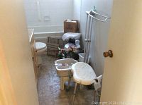 Photo showing bathroom doorway view with shower chair, foot bath tub, crutches, cleaning bottles, floral decor, and cardboard box.