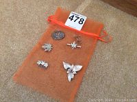 Five sterling silver brooches placed on an orange mesh pouch against carpet background; includes maple leaf and floral motifs.